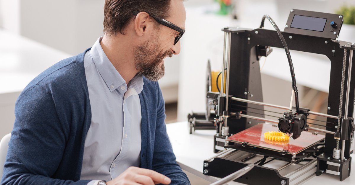 Hombre observando una impresora 3D mientras fabrica una pieza mecánica en plástico amarillo